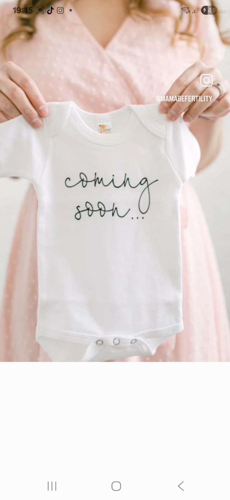 A small baby suit reading "coming soon..." — held in front of a pink dress.